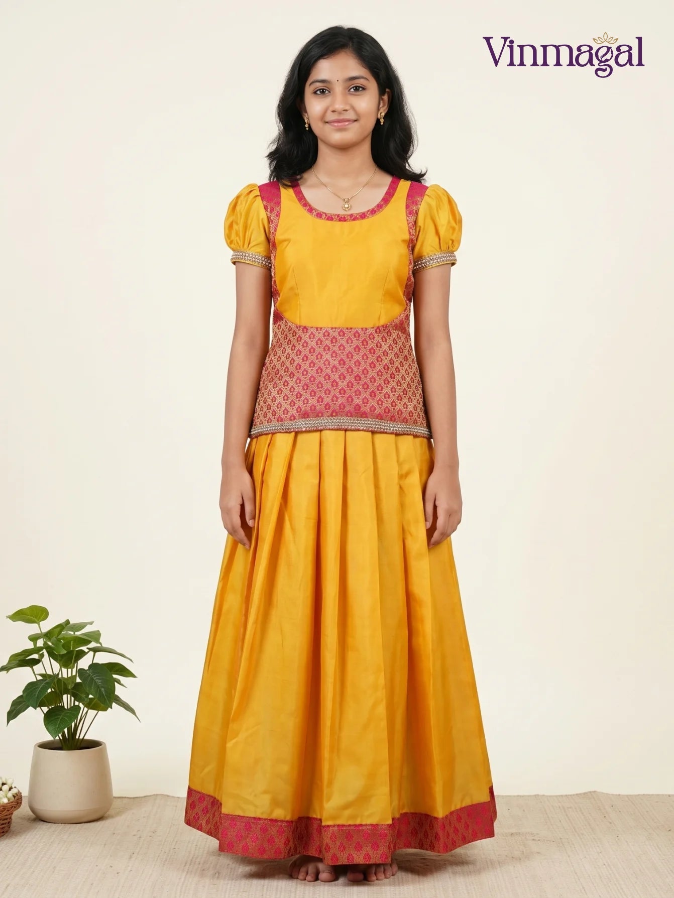 Traditional girls' mustard yellow cotton pavadai sattai from Vinmagal featuring a pink brocade waist panel and matching borders on the puff sleeves and pleated skirt hem.