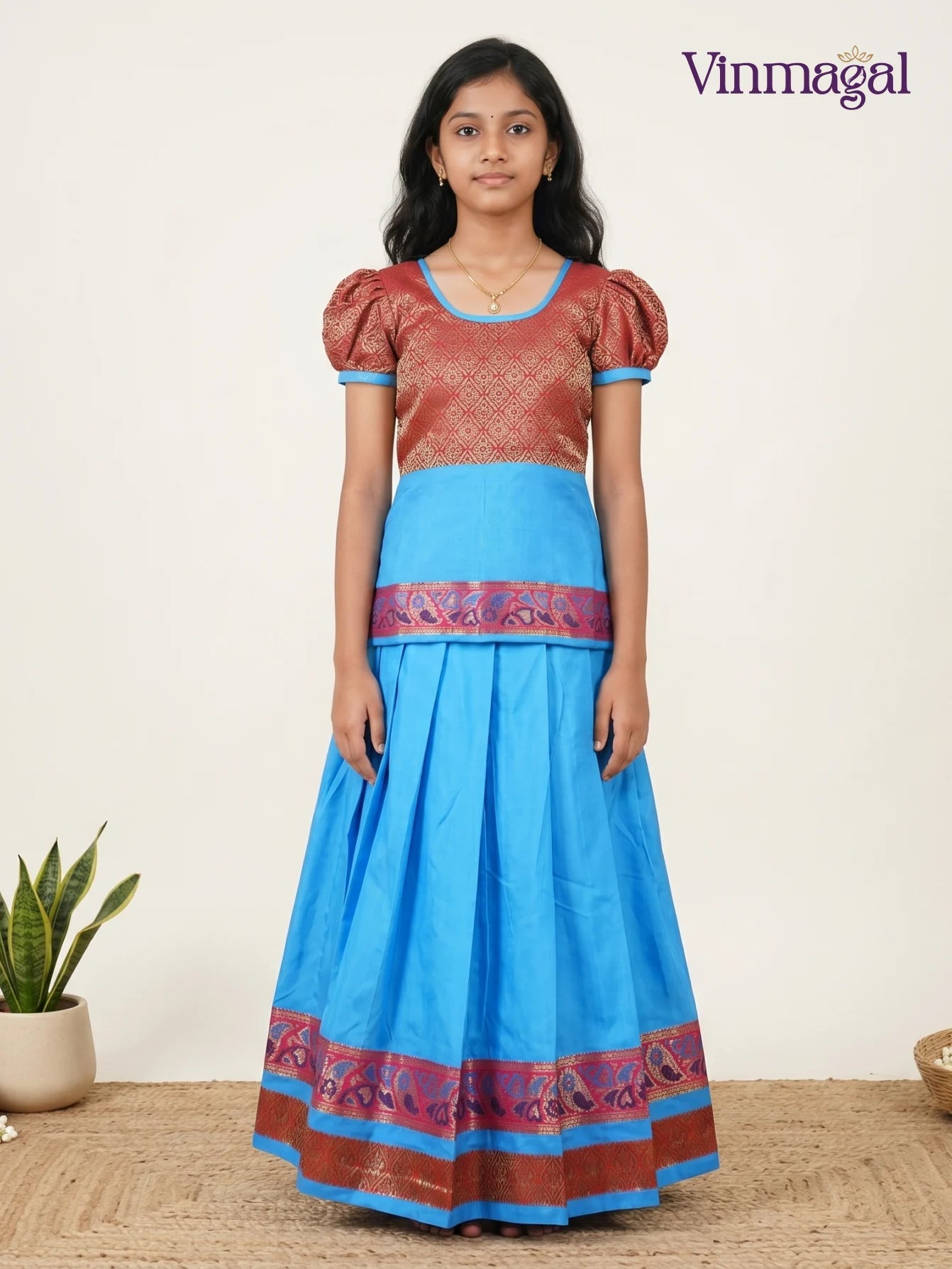 A young girl wearing a Vinmagal traditional cotton pavadai sattai set featuring a maroon brocade puff-sleeve top and a pleated sky-blue skirt with intricate ethnic borders.